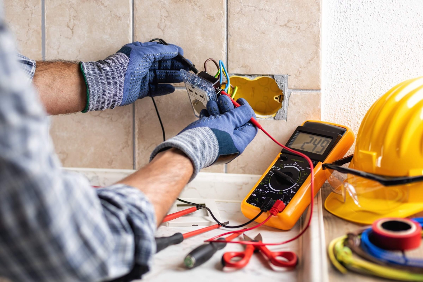 Electrician wiring a wall outlet with a multimeter and hard hat nearby